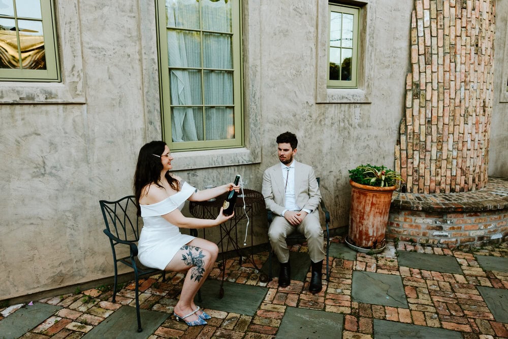 bride popping bottle while couple sits at small table against wall