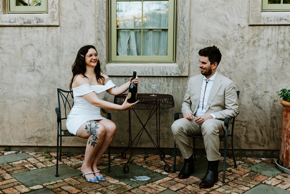 bride popping bottle while couple sits at small table against wall