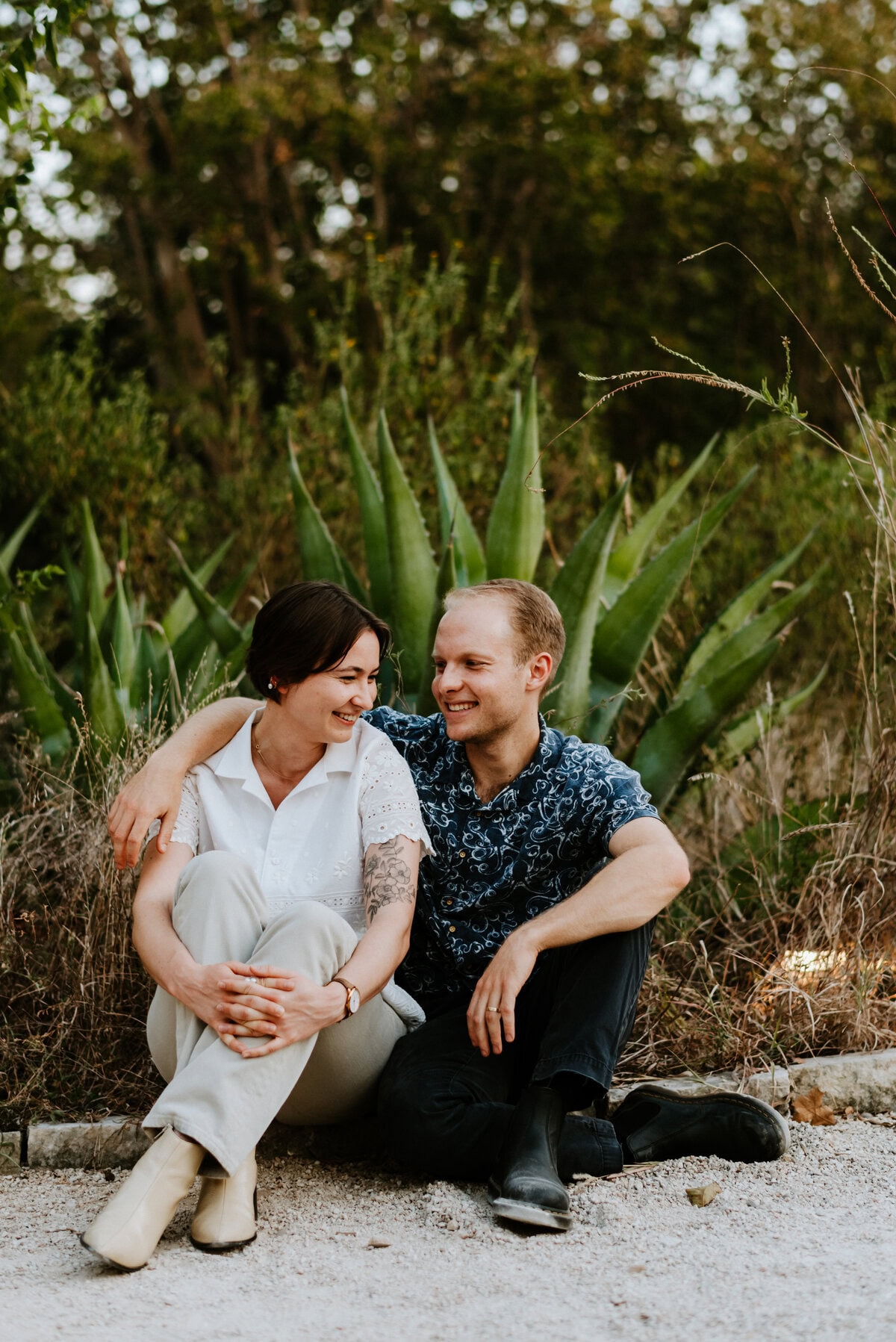 couple sitting on ground in front of greenery laughing