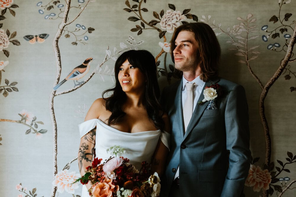 bride and groom stand close looking to the side with dynamic lighting in front of vintage wallpaper