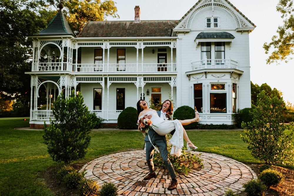 wide angle photo of groom holding bride in arms both smiling with joy