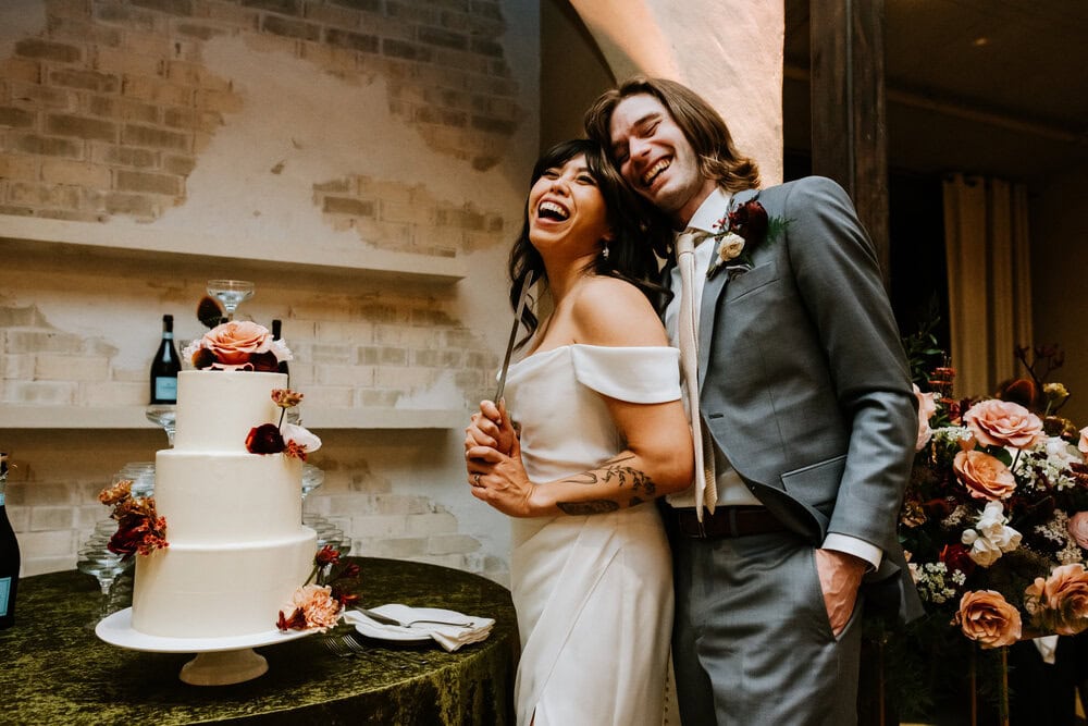 couple stands at cake table holding the knife smiling big