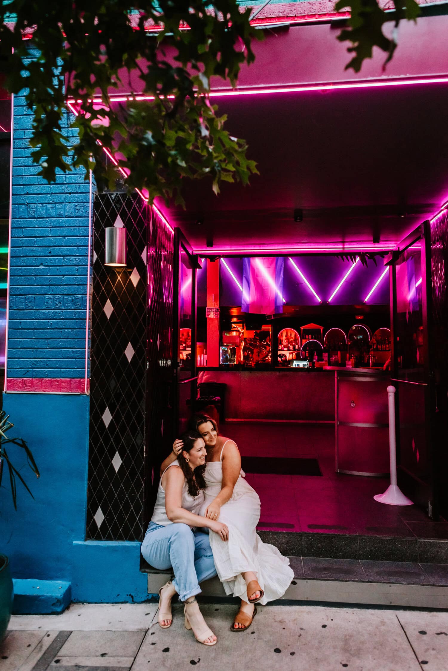 lesbian couple leans into each other while sitting in front of neon lights at restaurant 
