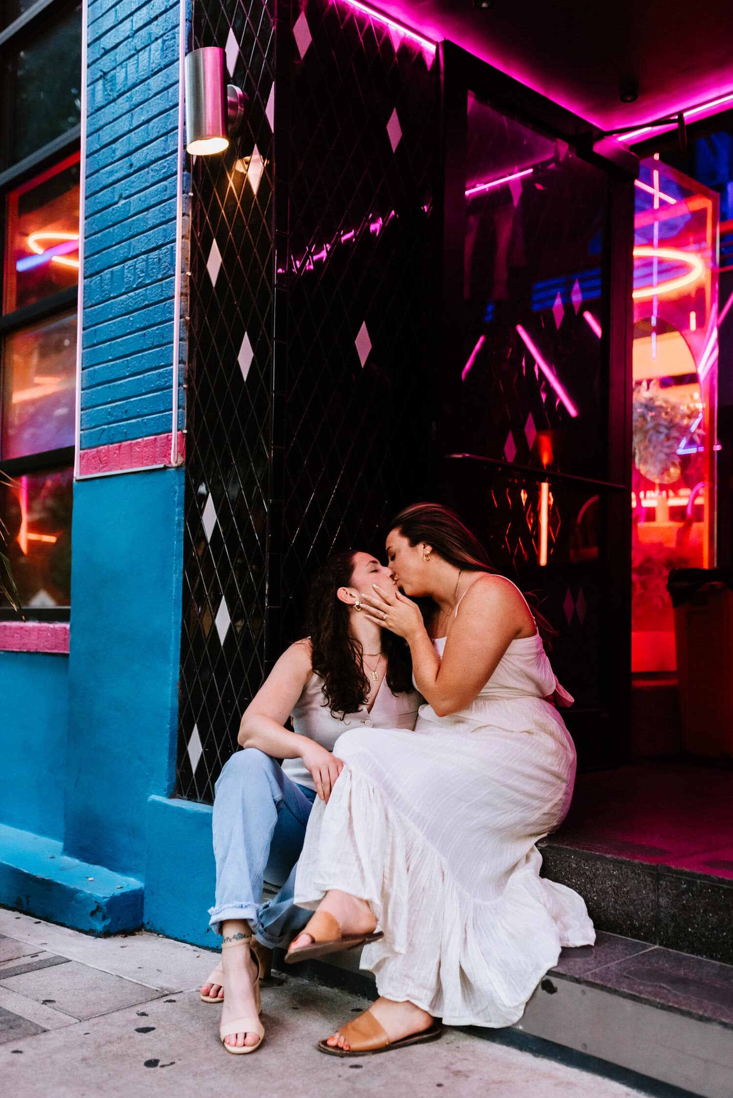 lesbian couple kisses each other while sitting in front of neon lights at restaurant 